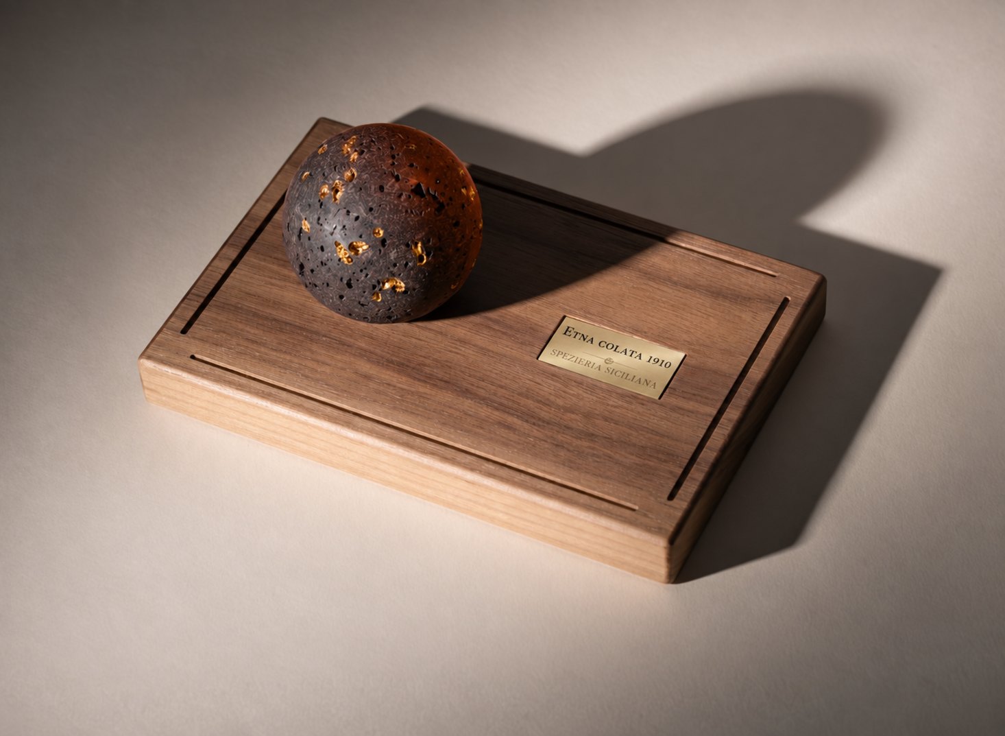 La sfera in lava dell'Etna sulla base in noce canaletto, con la targhetta «Etna colata 1910». Testi nell'immagine: «Un profumo per abitare lo spazio più intimo della casa.» «Base in legno di noce canaletto massello. Sfera in lava dell'Etna “occhio di pernice”.» «Poche gocce sui fori: la lava le trattiene e poi le rilascia lentamente.» «Trasforma il tempo in un momento.»